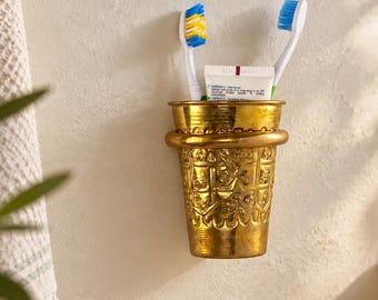 May include: A wall-mounted gold-toned metal cup featuring an embossed pattern of repeating geometric shapes. The cup holds two toothbrushes, one with blue and yellow bristles and one with blue bristles, along with a small white tube of toothpaste.