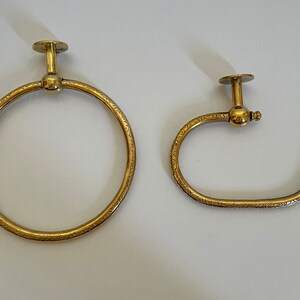 May include: Two brass-colored towel rings. One is a full circle, the other is a half-circle. Both have a decorative etched pattern and a mounting plate. The fixtures are designed for a bathroom.