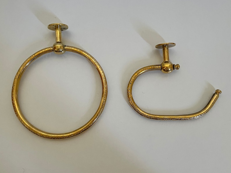 May include: Two brass-colored towel rings. One is a full circle, the other is a half-circle. Both have a decorative etched pattern and a mounting plate. The fixtures are designed for a bathroom.
