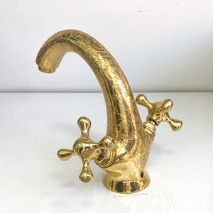 Op de afbeelding: Een gouden kraan met een decoratief, versierd ontwerp. De kraan heeft twee handvatten met een kruisvormig ontwerp.