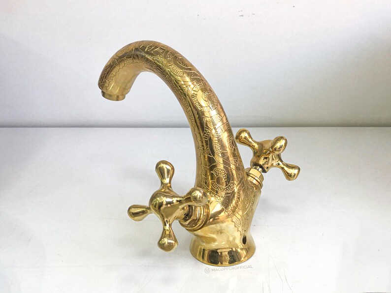 Op de afbeelding: Een gouden kraan met een decoratief, versierd ontwerp. De kraan heeft twee handvatten met een kruisvormig ontwerp.