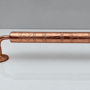 May include: A polished copper door handle featuring a cylindrical bar with a repeating embossed leaf pattern and a smooth, curved mounting bracket with a circular base plate containing a single screw hole for installation.