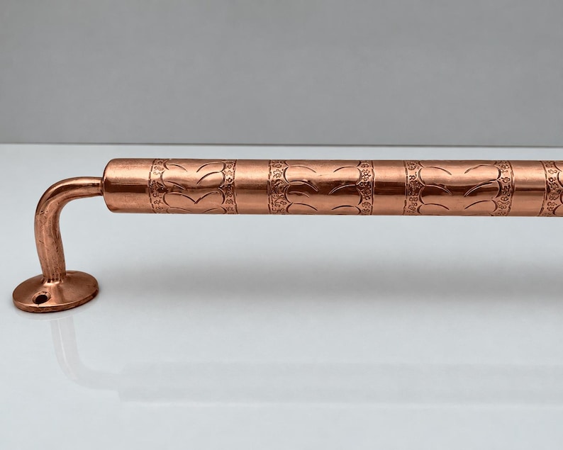 May include: A polished copper door handle featuring a cylindrical bar with a repeating embossed leaf pattern and a smooth, curved mounting bracket with a circular base plate containing a single screw hole for installation.