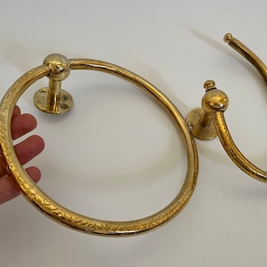 May include: Two brass towel rings. One is a circular ring with an etched floral pattern, and the other is a curved bar. Both have a round mounting plate and a ball-shaped detail.