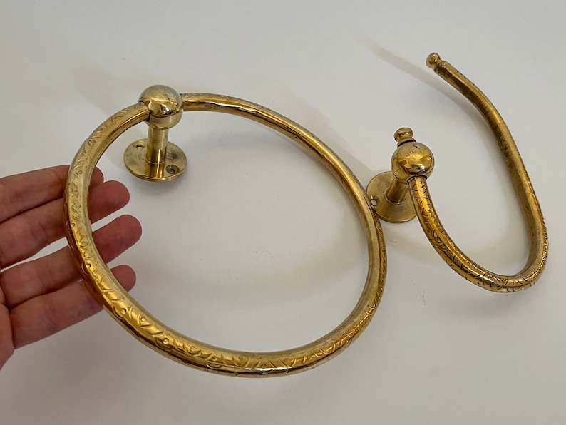 May include: Two brass towel rings. One is a circular ring with an etched floral pattern, and the other is a curved bar. Both have a round mounting plate and a ball-shaped detail.