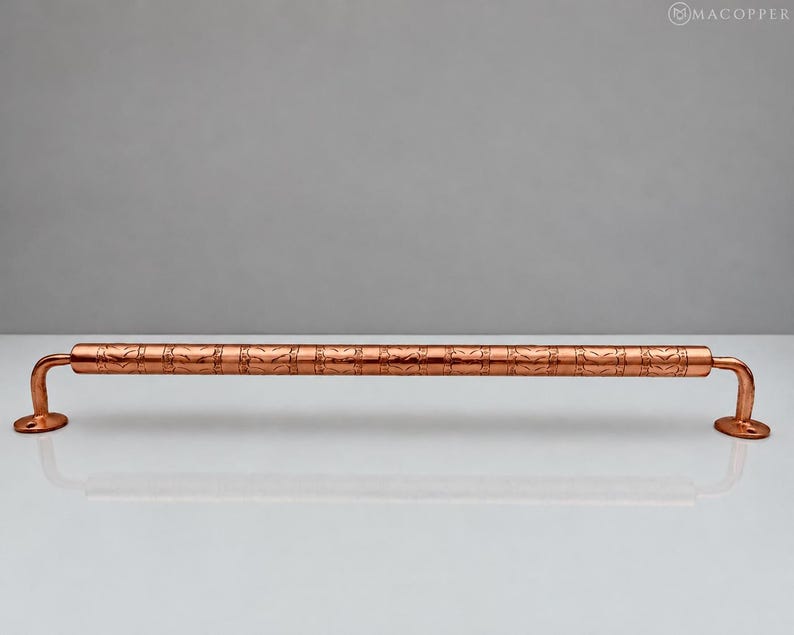 May include: A horizontal copper towel bar featuring a cylindrical rod with a repeating etched chevron pattern. The bar is supported by two curved mounting brackets on each end, each with a circular base plate containing a screw hole.
