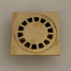 May include: A square, gold-colored drain cover with a circular grate. The grate has a series of small, rectangular openings.