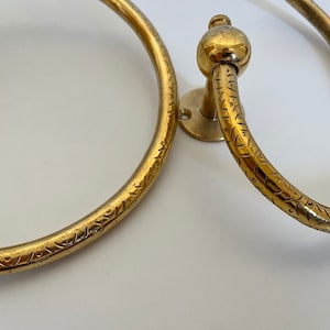 May include: Two antique brass towel rings. The rings have a circular design with decorative etched patterns. One ring has a ball-shaped detail. The rings are mounted on small circular plates.