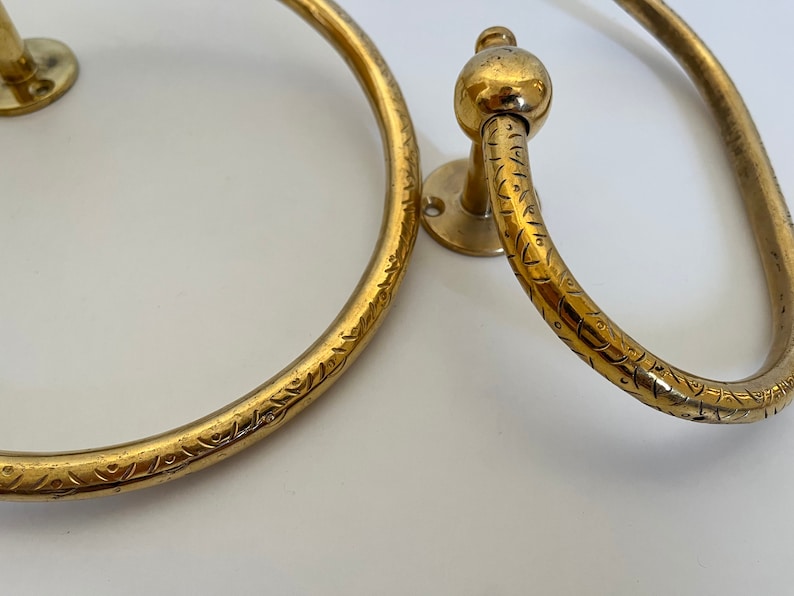 May include: Two antique brass towel rings. The rings have a circular design with decorative etched patterns. One ring has a ball-shaped detail. The rings are mounted on small circular plates.