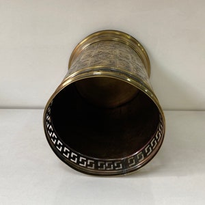 May include: A gold metal waste bin with a decorative pattern around the top edge. The inside of the bin is black.