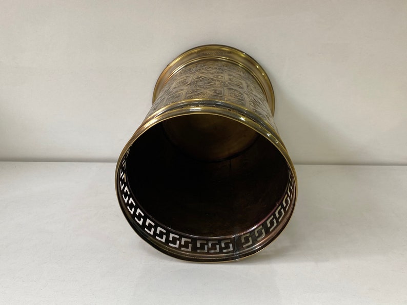 May include: A gold metal waste bin with a decorative pattern around the top edge. The inside of the bin is black.