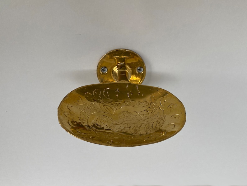May include: A gold-colored soap dish with an ornate design. The dish is mounted to a wall with two screws.