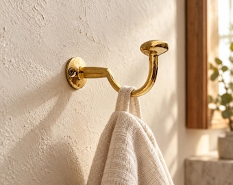 May include: A polished gold-toned metal wall hook with a rounded knob tip, mounted to a textured wall with a single visible screw. A beige, woven fabric towel hangs from the curved arm of the hook, creating a simple and functional storage display.