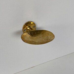 May include: A gold-toned door handle with an ornate, engraved design. The handle is shaped like a shallow bowl.