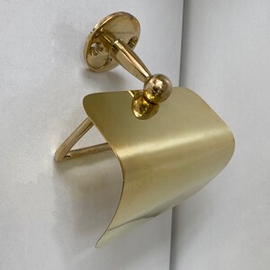 May include: A gold-toned metal toilet roll holder with a curved, flat design. The holder is mounted to a wall with a round base and a decorative knob.
