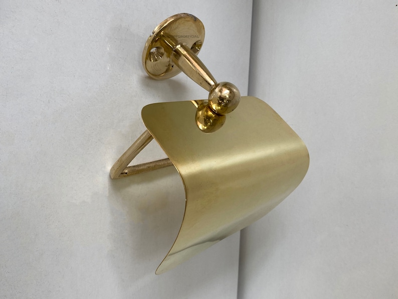 May include: A gold-toned metal toilet roll holder with a curved, flat design. The holder is mounted to a wall with a round base and a decorative knob.