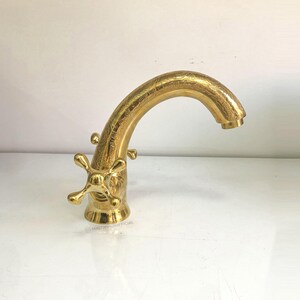 Op de afbeelding: Een gouden kraan met een gebogen uitloop en twee kruisvormige handvatten. De kraan heeft een getextureerde afwerking.
