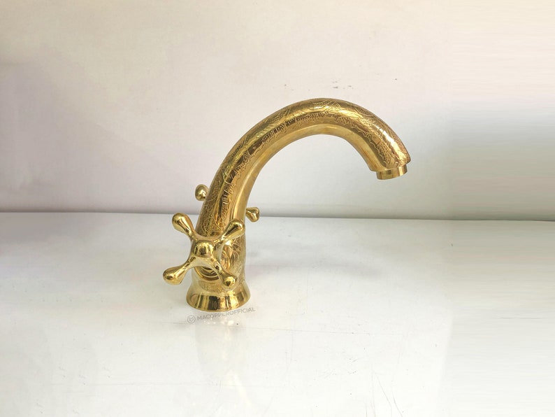 Op de afbeelding: Een gouden kraan met een gebogen uitloop en twee kruisvormige handvatten. De kraan heeft een getextureerde afwerking.