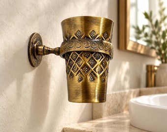 May include: A wall-mounted antique brass tumbler holder featuring a cylindrical cup with a textured diamond pattern and a decorative band. The metal fixture includes a circular base plate attached to the wall, supporting the cup holder.