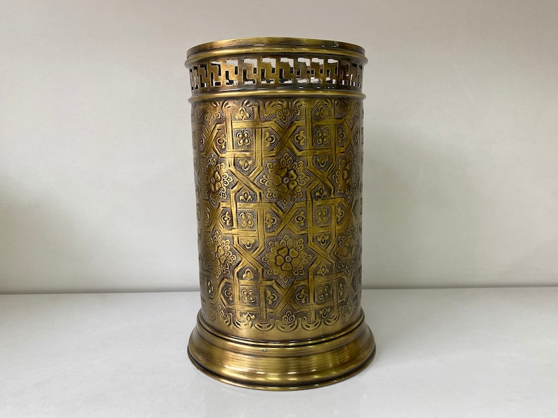 May include: A gold metal umbrella stand with an intricate geometric pattern. The stand has a round base and a cylindrical body.