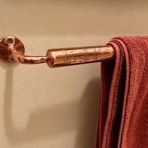May include: A polished copper towel bar mounted to a wall with two visible screws. The bar features a cylindrical section with an engraved pattern and holds a folded, solid dark red towel draped over the horizontal metal rod.