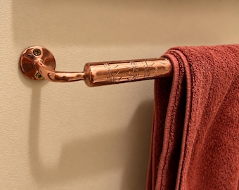Puede incluir: Un toallero de cobre pulido montado en la pared con dos tornillos visibles. La barra presenta una sección cilíndrica con un patrón grabado y sostiene una toalla de color rojo oscuro sólido doblada sobre la varilla.