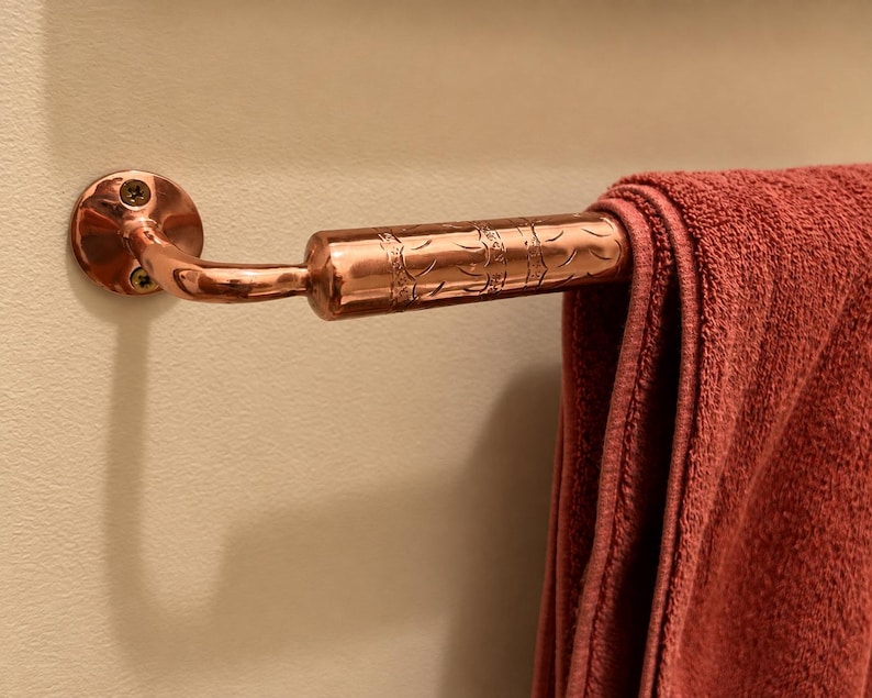 May include: A polished copper towel bar mounted to a wall with two visible screws. The bar features a cylindrical section with an engraved pattern and holds a folded, solid dark red towel draped over the horizontal metal rod.