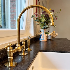 May include: A brass kitchen faucet with a high-arched spout and two side handles, featuring a weathered metallic finish. The faucet is mounted on a dark stone countertop next to a white ceramic sink basin, with a decorative vase nearby.