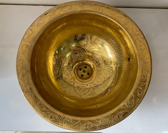 Può includere: Un lavandino in metallo dorato con un design ornamentale attorno al bordo. Il lavandino ha uno scarico al centro.