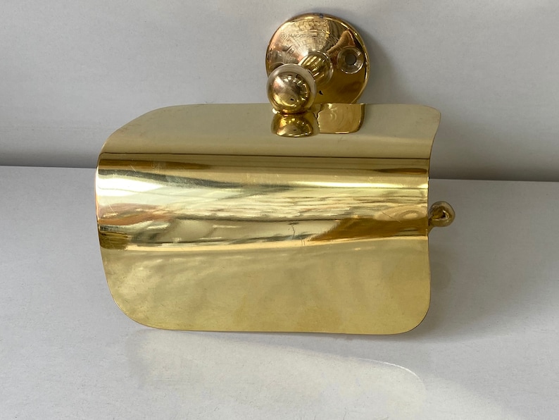 May include: A gold-toned toilet roll holder with a curved design. The holder is mounted to a wall.
