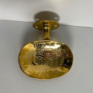 May include: A gold-toned metal door handle with an oval-shaped plate featuring an intricate floral design.