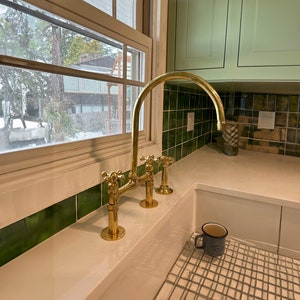 May include: A gold kitchen tap with a high arc spout and cross handles. The tap is installed in a white kitchen sink with green tiled backsplash.