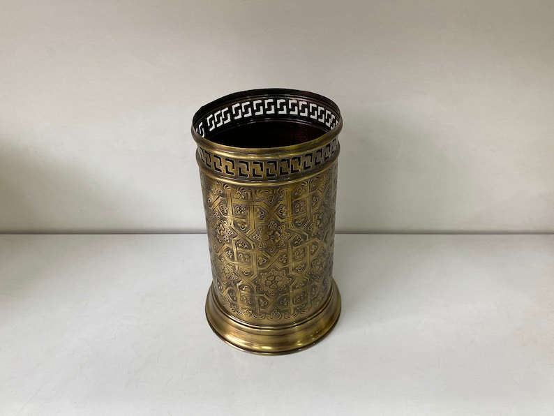 May include: A gold metal umbrella stand with an intricate geometric pattern. The stand has a decorative border around the top.