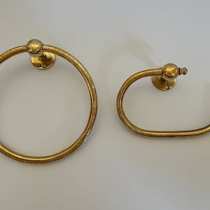 May include: Two antique brass bathroom fixtures: a towel ring and a toilet paper holder. Both have a circular design with etched detailing and a ball-shaped accent. The fixtures are mounted on a white surface.