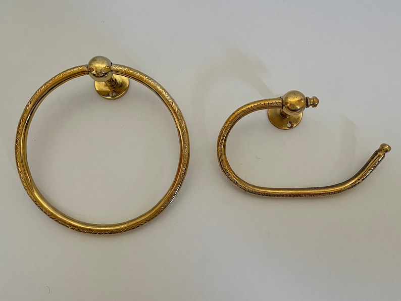 May include: Two antique brass bathroom fixtures: a towel ring and a toilet paper holder. Both have a circular design with etched detailing and a ball-shaped accent. The fixtures are mounted on a white surface.