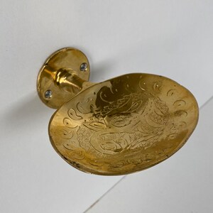 May include: A gold-toned soap dish with an ornate design. The dish is attached to a wall mount.