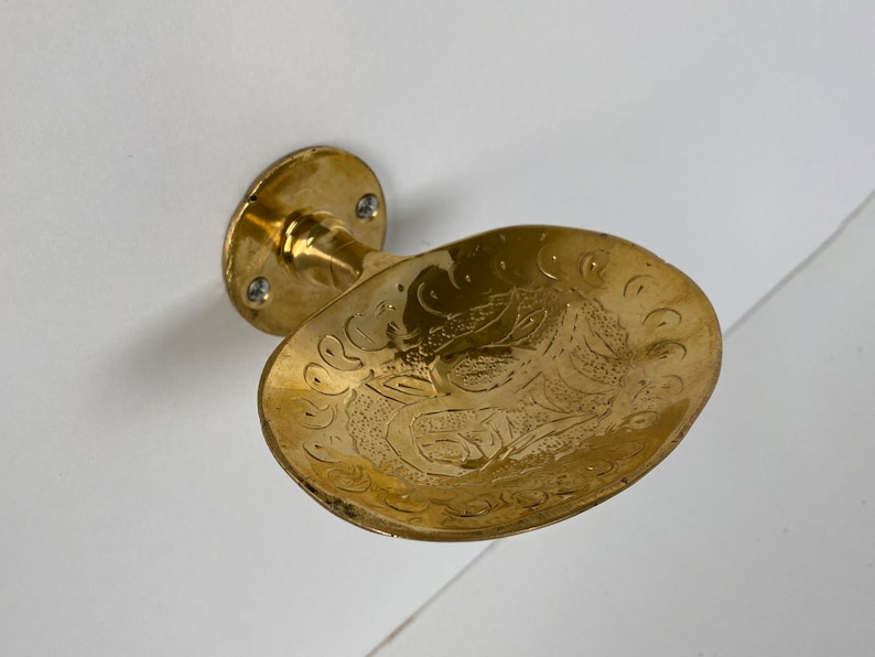 May include: A gold-toned soap dish with an ornate design. The dish is attached to a wall mount.