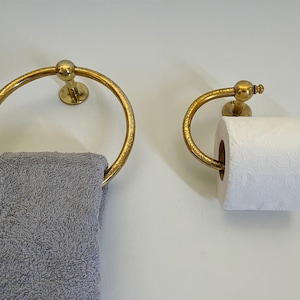 May include: A gold-colored towel ring and toilet paper holder set. The towel ring is circular, and the toilet paper holder has a curved design. Both are mounted on a white wall. A gray towel hangs from the ring.
