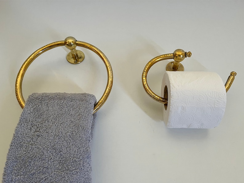 May include: A gold-colored towel ring and toilet paper holder set. The towel ring is circular, and the toilet paper holder has a curved design. Both are mounted on a white wall. A gray towel hangs from the ring.