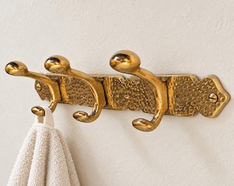 May include: A brass wall-mounted coat rack featuring three double hooks attached to a rectangular backplate with a hammered texture. The rack is secured with two visible screws and holds a beige textured towel on the leftmost hook.