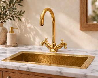 May include: A rectangular hammered gold sink basin set into a white marble countertop with a high-arched gold faucet featuring two cross-handle knobs. The sink has a textured, dimpled surface finish and a polished metallic gold appearance.