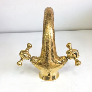 Op de afbeelding: Een gouden kraan met ingewikkelde gegraveerde ontwerpen. De kraan heeft twee handvatten met een kruisvormig ontwerp.