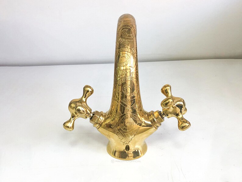 Op de afbeelding: Een gouden kraan met ingewikkelde gegraveerde ontwerpen. De kraan heeft twee handvatten met een kruisvormig ontwerp.