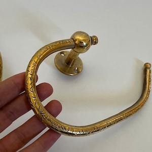 May include: A pair of antique brass towel rings. The rings have a circular design with an etched pattern. One ring is mounted on a round base. The brass has a warm, aged patina, suggesting vintage or antique origins. The rings are designed for bathroom use.