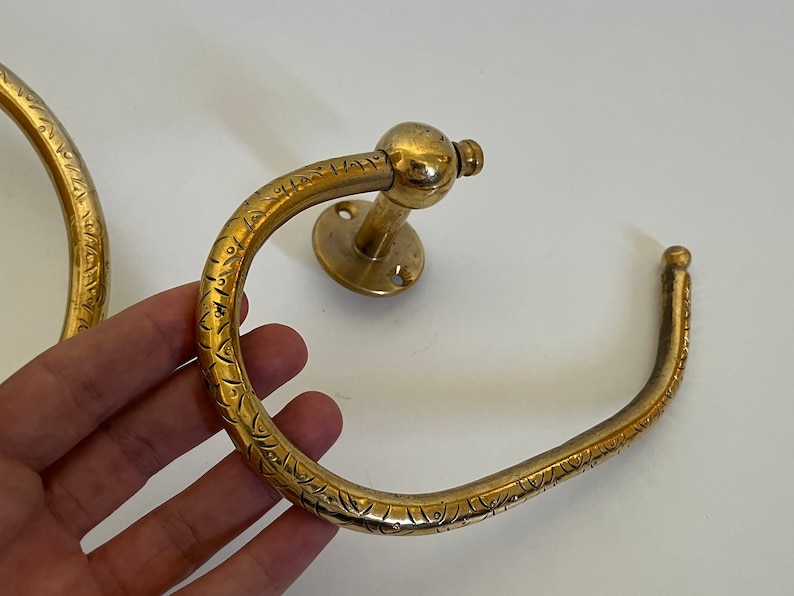 May include: A pair of antique brass towel rings. The rings have a circular design with an etched pattern. One ring is mounted on a round base. The brass has a warm, aged patina, suggesting vintage or antique origins. The rings are designed for bathroom use.