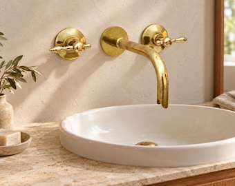 Puede incluir: Un grifo de pared de latón con dos manijas de palanca a juego situado sobre un lavabo de cerámica blanca. El grifo presenta un caño curvo y un acabado metálico pulido, proporcionando un accesorio funcional para un mueble de baño.