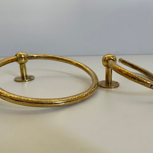 May include: Two brass towel rings. The larger ring is approximately 8 inches in diameter, and the smaller ring is about 4 inches in diameter. Both rings have a decorative etched pattern and are mounted on a small, round base.
