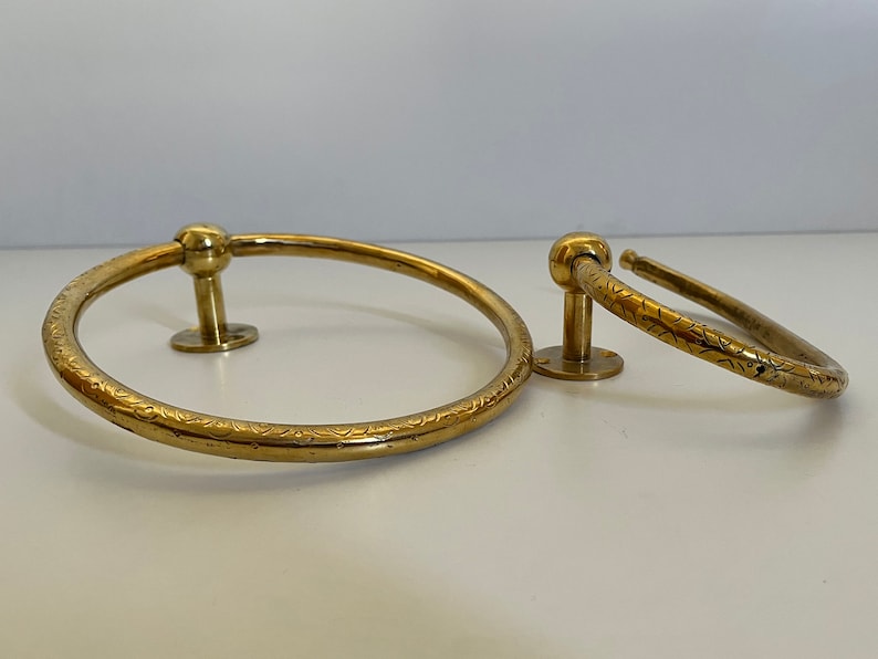 May include: Two brass towel rings. The larger ring is approximately 8 inches in diameter, and the smaller ring is about 4 inches in diameter. Both rings have a decorative etched pattern and are mounted on a small, round base.