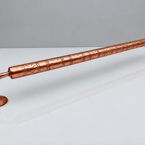 May include: A polished copper grab bar featuring a cylindrical rod with a repeating embossed leaf pattern. The bar is supported by two curved brackets, each ending in a circular mounting plate with pre-drilled holes for installation.