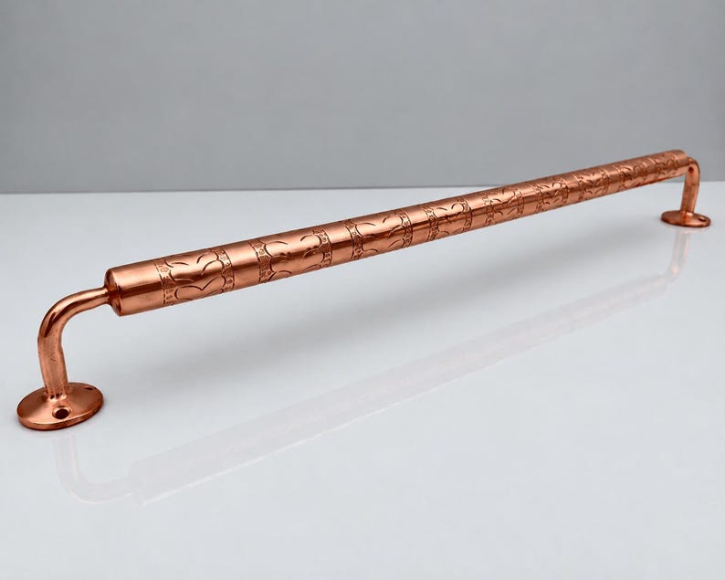 May include: A polished copper grab bar featuring a cylindrical rod with a repeating embossed leaf pattern. The bar is supported by two curved brackets, each ending in a circular mounting plate with pre-drilled holes for installation.